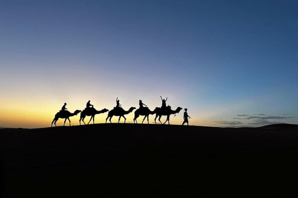 Silhouette of travelers camel trekking across the Merzouga Sahara Desert at sunset during a private Morocco desert tour.